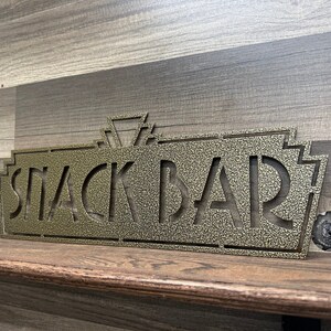 May include: A gold metal sign with an art deco design that reads "Snack Bar".