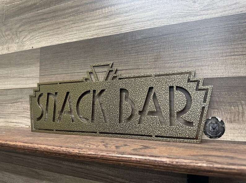 May include: A gold metal sign with an art deco design that reads "Snack Bar".
