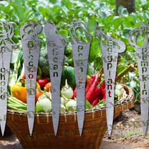 May include: Six metal garden markers with decorative tops and plant names: Artichoke, Broccoli, Bean, Pepper, Eggplant, Pumpkin. The markers are in a basket with various vegetables.