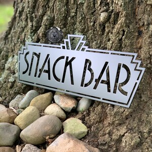 May include: A silver metal sign with a vintage art deco design that reads "Snack Bar".