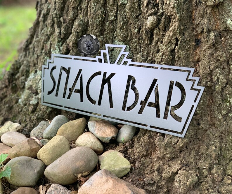 May include: A silver metal sign with a vintage art deco design that reads "Snack Bar".