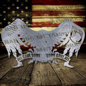 Può includere: Un ritaglio di metallo di un'aquila con le ali spiegate. L'aquila tiene uno striscione che recita "All Gave Some, Some Gave All". L'aquila tiene anche un simbolo POW/MIA. Il ritaglio è su uno sfondo di legno con una bandiera americana sullo sfondo.