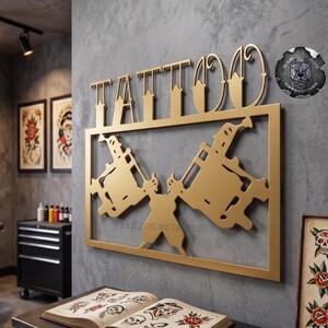 May include: A gold-colored metal wall sign featuring the word TATTOO in stylized lettering above a rectangular frame containing two crossed tattoo machines. A circular logo with a dog illustration and the text BAD DOG METALWORKS is visible.