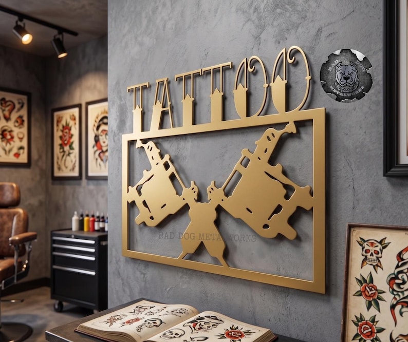 May include: A gold-colored metal wall sign featuring the word TATTOO in stylized lettering above a rectangular frame containing two crossed tattoo machines. A circular logo with a dog illustration and the text BAD DOG METALWORKS is visible.