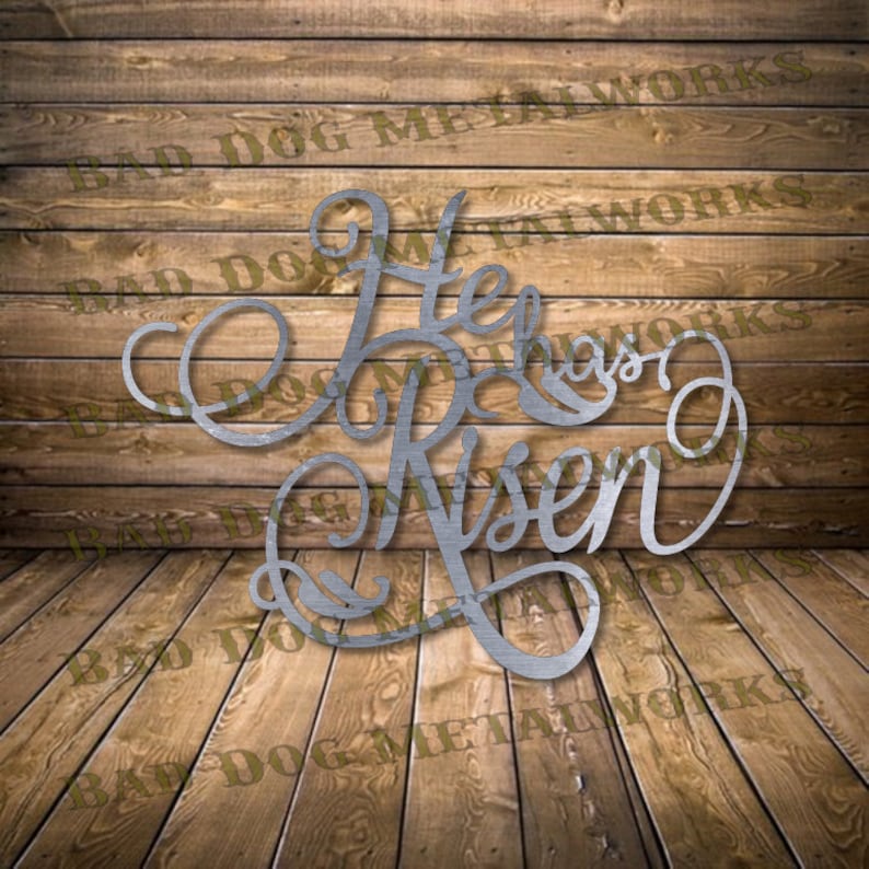 May include: Metal wall art with a cursive script design that reads "He Has Risen". The art is silver in color and is displayed against a wood background.
