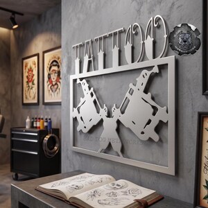 May include: A silver-colored metal wall sign featuring the word TATTOO in stylized lettering above a rectangular frame containing two crossed tattoo machines. A circular logo with the text BAD DOG METALWORKS is visible in the upper right corner.
