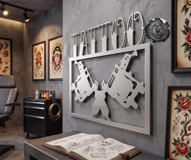 May include: A silver-colored metal wall sign featuring the word TATTOO in stylized lettering above a rectangular frame containing two crossed tattoo machines. A circular logo with the text BAD DOG METALWORKS is visible in the upper right corner.