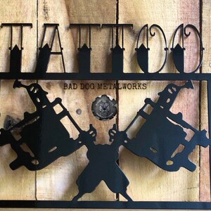 May include: A black metal wall sign featuring the word TATTOO in large capital letters at the top. Below, two crossed tattoo machines are framed within a rectangular border. The text BAD DOG METALWORKS and a circular logo are centered.