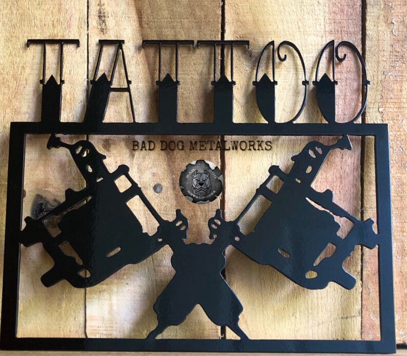 May include: A black metal wall sign featuring the word TATTOO in large capital letters at the top. Below, two crossed tattoo machines are framed within a rectangular border. The text BAD DOG METALWORKS and a circular logo are centered.
