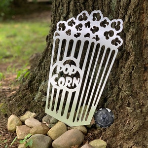 May include: A metal cutout of a popcorn bucket with the word "POPCORN" on the front. The cutout is silver and has a rustic look. It is sitting on a tree trunk with rocks around it.