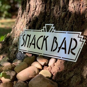 May include: A metal sign with a vintage art deco design that reads "Snack Bar". The sign is leaning against a tree trunk.