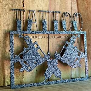 May include: A grey metal sign with a speckled finish featuring the word TATTOO in large capital letters at the top. Below the text, the sign displays two crossed tattoo machines and the text BAD DOG METALWORKS centered in the rectangular frame.