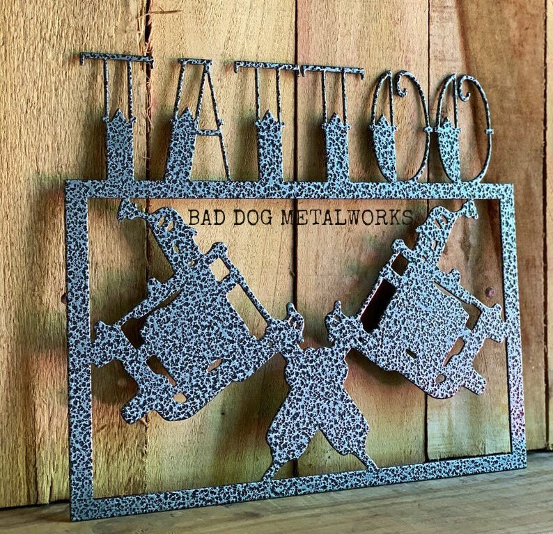 May include: A grey metal sign with a speckled finish featuring the word TATTOO in large capital letters at the top. Below the text, the sign displays two crossed tattoo machines and the text BAD DOG METALWORKS centered in the rectangular frame.