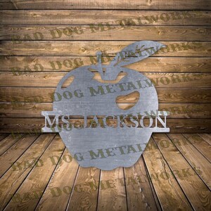 May include: A silver metal apple with a stem and leaf, with the name "MS. JACKSON" written across the middle. The apple is on a wood background.