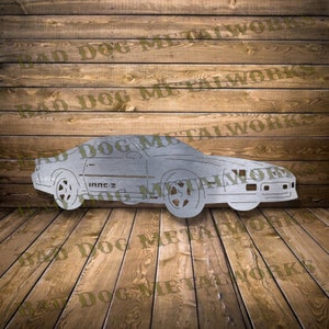 May include: A silver metal cutout of a 1980s Chevrolet Camaro IROC-Z. The car is facing to the right and has a detailed profile. The cutout is on a wooden background.