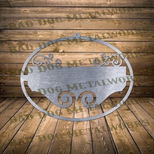 Pode incluir: Um sinal oval de metal prateado com um espaço retangular em branco no centro. O sinal tem detalhes decorativos em rolagem na parte superior e inferior. O texto "Bad Dog Metalworks" é visível no sinal.