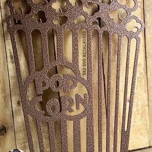 May include: A metal popcorn bucket cutout with the word "POPCORN" in the center. The bucket is brown and has a textured finish. The text "BAD DOG METALWORKS" is on the side of the bucket.