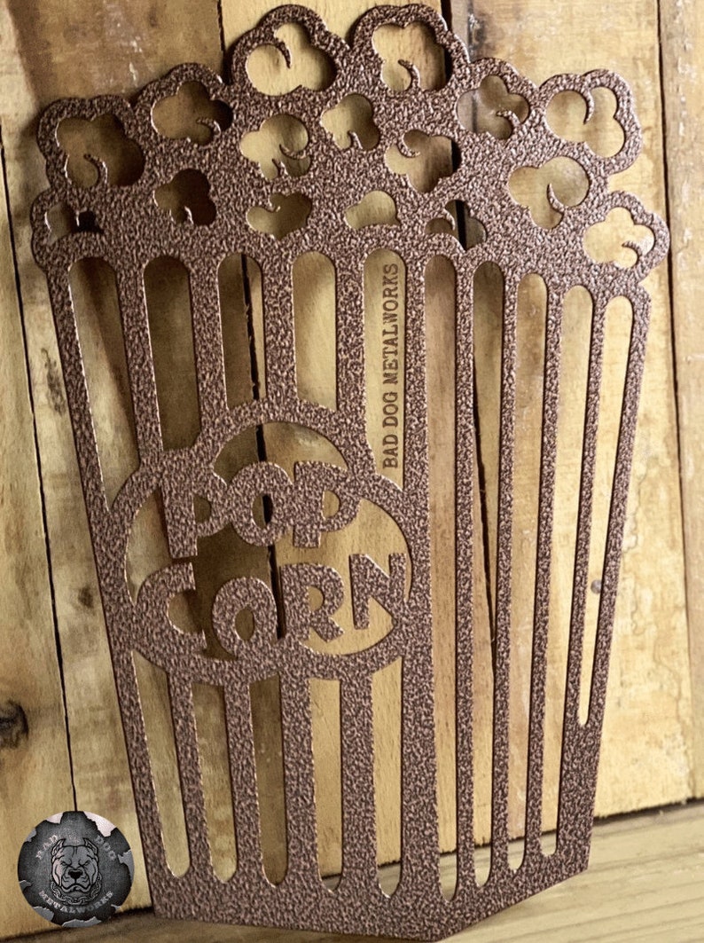 May include: A metal popcorn bucket cutout with the word "POPCORN" in the center. The bucket is brown and has a textured finish. The text "BAD DOG METALWORKS" is on the side of the bucket.