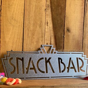 May include: A silver metal sign with the words "Snack Bar" in a retro art deco style. The sign is on a wooden background with candy and chocolate bars in the foreground.