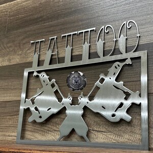 May include: A brushed metal wall sign featuring the word TATTOO in a stylized font above a rectangular frame. Inside the frame are two symmetrical tattoo machine silhouettes facing outward, with a circular logo in the center depicting a dog head.