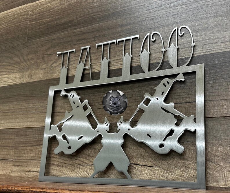 May include: A brushed metal wall sign featuring the word TATTOO in a stylized font above a rectangular frame. Inside the frame are two symmetrical tattoo machine silhouettes facing outward, with a circular logo in the center depicting a dog head.