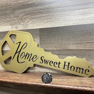 May include: Gold metal key-shaped sign with the words "Home Sweet Home" cut out. The sign is on a wood surface.