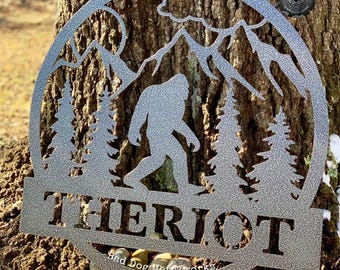 May include: A circular silver metal wall sign featuring a cutout silhouette of a Bigfoot walking through a forest with mountains in the background. The name THERIOT is cut out in bold capital letters across the bottom, with Bad Dog Metalworks text.