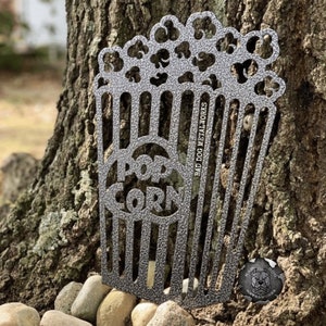 May include: A metal cutout of a popcorn box with the words "Pop Corn" inside. The cutout is a light gray color and has a textured surface. The cutout is sitting on a tree trunk with rocks around it.