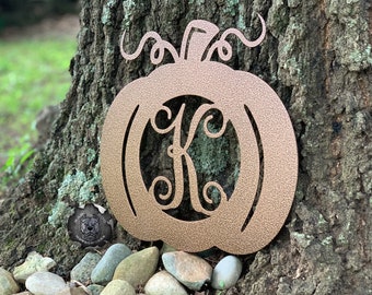 May include: A gold metal pumpkin with a decorative swirl design and a monogrammed letter "K" in the center. The pumpkin is sitting on a bed of rocks.