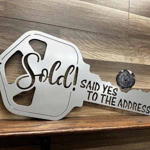 May include: A silver metal key-shaped sign with the word "Sold!" and the phrase "Said Yes To The Address" cut out. The sign is displayed on a wooden surface. A small, round emblem with a dog's face is next to the key.