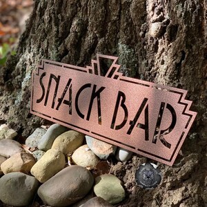 May include: A copper-colored metal sign with an art deco design that reads "Snack Bar". The sign is sitting on a bed of small, smooth rocks.