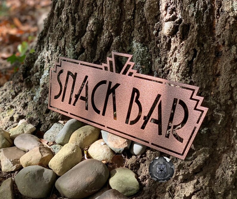 May include: A copper-colored metal sign with an art deco design that reads "Snack Bar". The sign is sitting on a bed of small, smooth rocks.