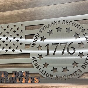 May include: A silver metal American flag with a circle in the center that says "When Tyranny Becomes Law Rebellion Becomes Duty" and "1776" inside the circle. The flag is on a wooden surface with a sign that says "Bad Dog Metalworks".