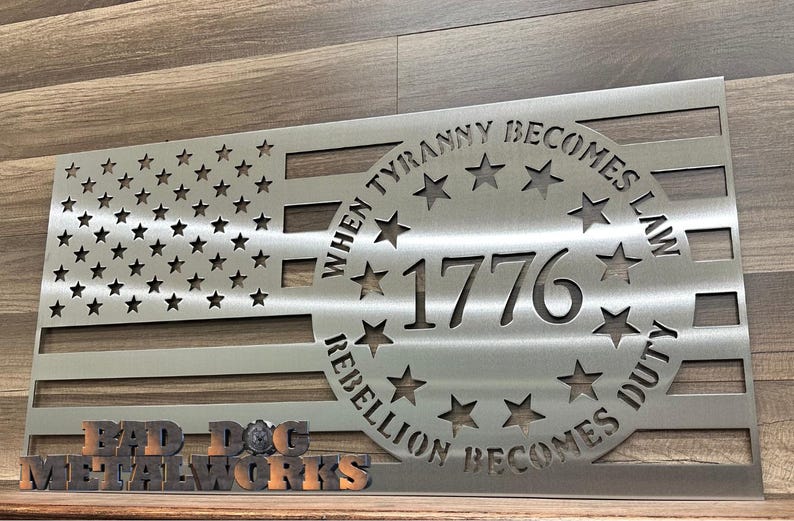 May include: A silver metal American flag with a circle in the center that says "When Tyranny Becomes Law Rebellion Becomes Duty" and "1776" inside the circle. The flag is on a wooden surface with a sign that says "Bad Dog Metalworks".