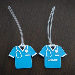 May include: Two blue medical scrubs-shaped luggage tags with white accents and a stethoscope design. One tag has the name "GRACE" in white. Each tag has a clear plastic loop for attachment. The tags are on a dark brown wooden surface.