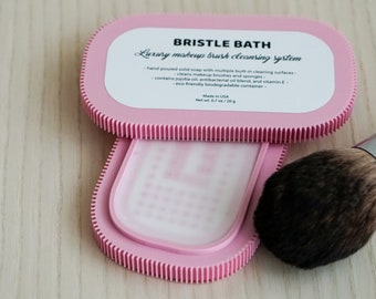 May include: A pink plastic container with the text "BRISTLE BATH Luxury makeup brush cleaning system" and a white bar of soap inside. The container is open and a makeup brush is lying next to it.