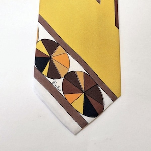Op de afbeelding: Een vintage stropdas met een geometrisch ontwerp. De stropdas heeft een gele achtergrond met bruine en oranje vormen. Het onderste gedeelte heeft een witte rand met cirkelvormige patronen in bruin, oranje en zwart. De stropdas is gesigneerd "Fulvio".