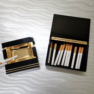 Pode incluir: Um cinzeiro preto e dourado com um cigarro e um porta-cigarros preto com cigarros. O cinzeiro tem um interior dourado e um exterior preto com acabamentos dourados. O porta-cigarros está aberto, revelando cigarros.