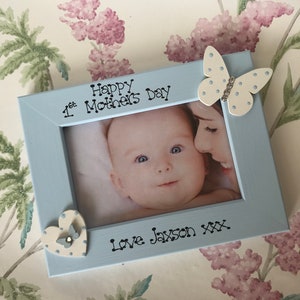May include: A pale blue picture frame with a photograph of a baby and a woman. The frame is decorated with a butterfly and heart. The text on the frame reads "Happy Mother's Day" and "Love Jayson xxx".