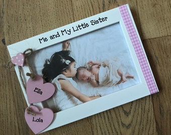 May include: White photo frame with a pink and white gingham ribbon. The frame has a photo of two sisters, one older and one younger. The frame has two pink wooden hearts with the names "Ella" and "Lola" written on them. The frame has the text "Me and My Little Sister" written on it.