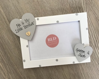 May include: White rectangular picture frame with gray polka dots. Two gray heart-shaped embellishments are attached, one with "Me & My Little Brother" and a button, the other with "George & Jacob" in black script.