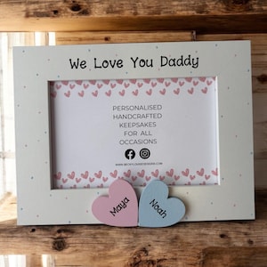 May include: White wooden picture frame with a white background and pink and blue hearts. The frame has the text "We Love You Daddy" at the top and the text "Maya" and "Noah" on two wooden hearts at the bottom.