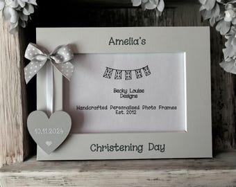 May include: A gray wooden photo frame with a white mat and a heart-shaped wooden ornament. The frame is personalized with the name "Amelia's" and the date "10.11.2024". The text "Christening Day" is also included on the frame.