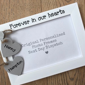May include: White photo frame with the text "Forever in our hearts" on the top. The frame has two grey wooden hearts hanging from it with the names "Nanny" and "Hampton" written on them.