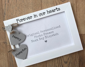 May include: White photo frame with the text "Forever in our hearts" on the top. The frame has two gray wooden hearts hanging from it with the names "Nanny" and "Hampton" written on them.