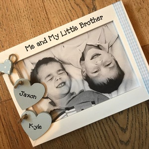 May include: White photo frame with a black and white photo of two boys. The frame has a blue and white gingham ribbon on the right side. The top of the frame says "Me and My Little Brother". There are three wooden hearts hanging from the top of the frame. The hearts are painted light blue and have the names "Jaxon" and "Kyle" written on them.