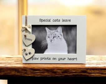 May include: A white picture frame with a black and white photo of a cat. The frame has the text "Special cats leave paw prints on your heart." Two heart-shaped ornaments are attached, one with the name "Poppy" and the other with paw prints.