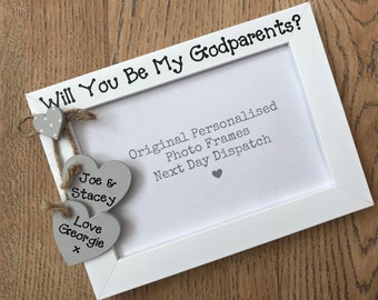 May include: A white rectangular photo frame featuring the text Will You Be My Godparents? at the top. Three grey wooden hearts are attached to the left side with twine. Two hearts are inscribed with Joe & Stacey and Love Georgie x.