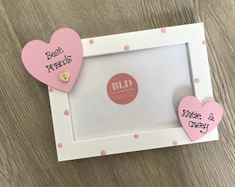 May include: White picture frame with pink polka dots and two pink wooden hearts. The hearts have the text "Best Friends" and "Katie & Casey" written on them.