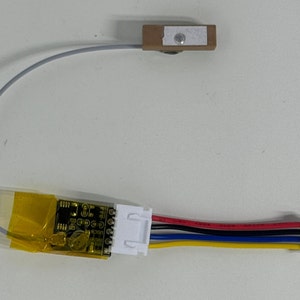 May include: A small circuit board with a yellow protective covering and a white connector with four wires: red, blue, black, and yellow. A gray wire connects to a small brown sensor with a silver button.